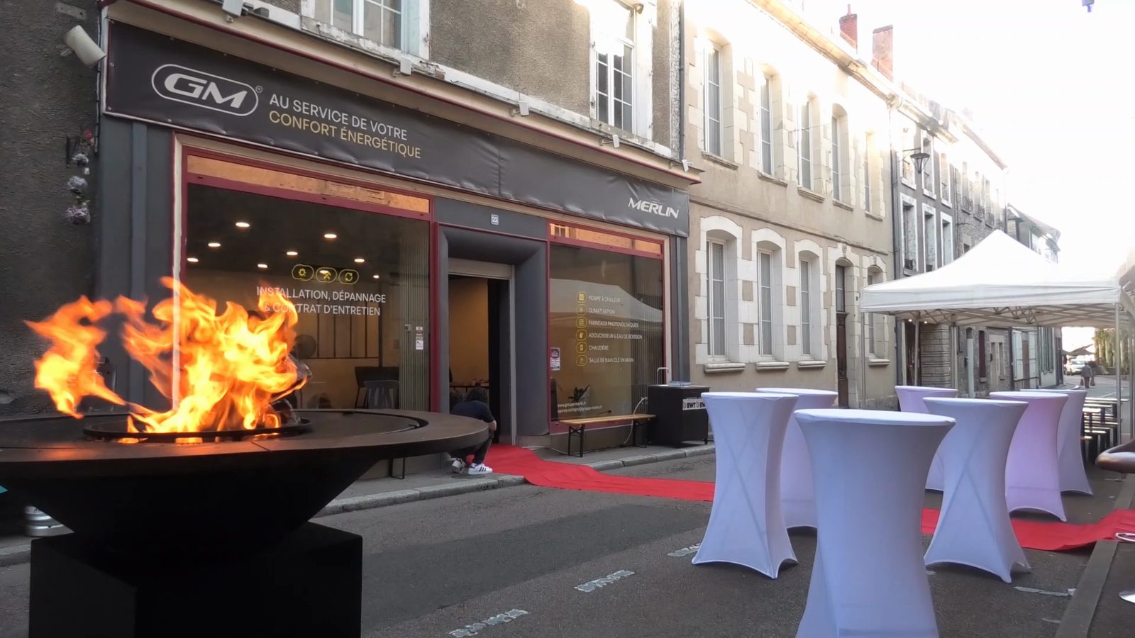 Brasero allumé devant la façade de l'agence Groupe Merlin de Corbigny, avec un tapis rouge et des tables hautes dressées dans la Grande Rue